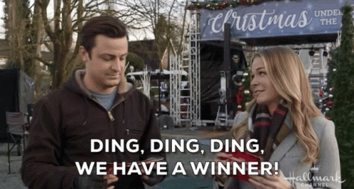 A Man And Woman Are Standing In Front Of A Sign That Says Christmas Under The Christmas Tree GIF
