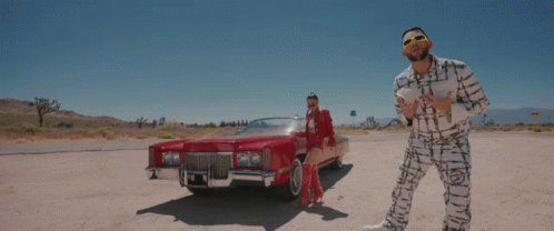 A Man And Woman Are Standing Next To A Red Car GIF