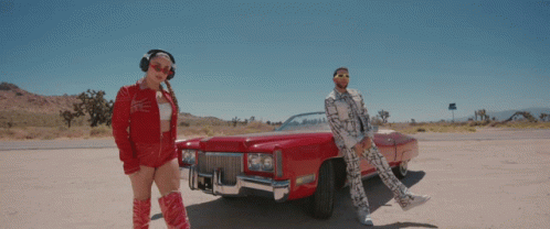 A Man And Woman Are Standing Next To A Red Car GIF