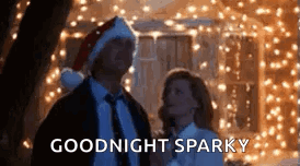 A Man And Woman Are Standing Next To Each Other In Front Of Christmas Lights And The Man Is Wearing A Santa Hat . GIF