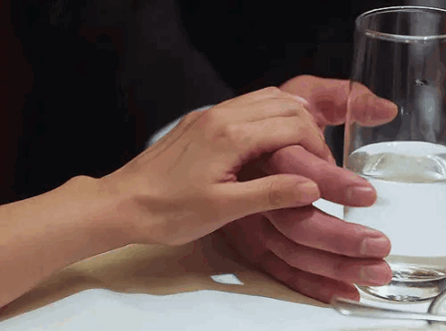 A Man And Woman Holding Hands Over A Glass Of Water On A Table GIF