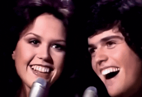 A Man And Woman Singing Into Microphones And Smiling GIF