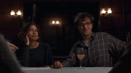 A Man And Woman Sitting At A Table With A Glass Of Wine GIF