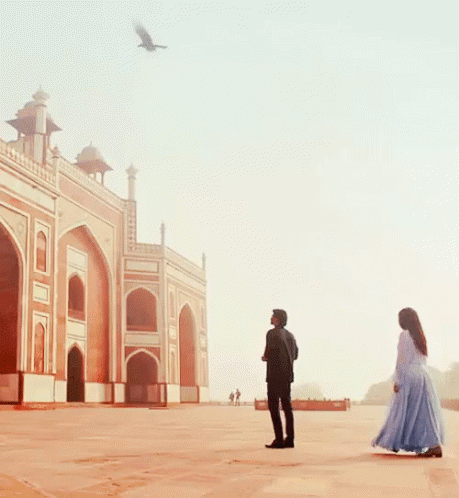 A Man And Woman Standing In Front Of A Building With A Bird Flying In The Sky GIF