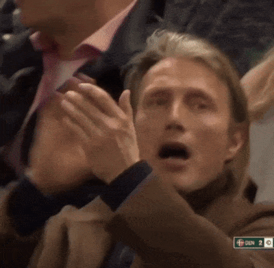 A Man Applauds While Sitting In A Crowd With A Sign That Says Den On It GIF