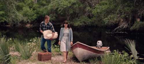 A Man Carrying A Blanket And A Woman Carrying A Picnic Basket Next To A Small Boat GIF