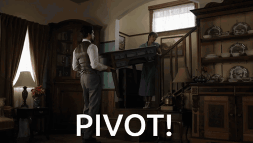 A Man Carrying A Table In A Living Room With The Word Pivot Below Him GIF
