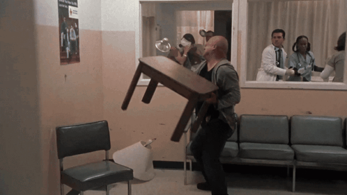 A Man Carrying A Table In A Waiting Room With A Red Cross Sign On The Wall GIF