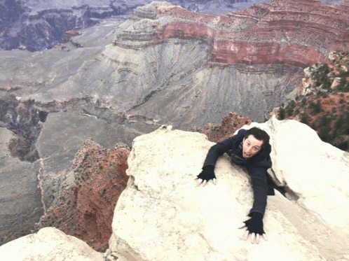 A Man Climbs Up A Rocky Cliff With A Valley In The Background GIF