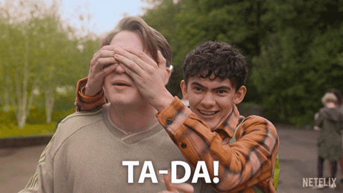 A Man Covering Another Man 'S Eyes With His Hands And The Words Ta-Da Are Visible GIF