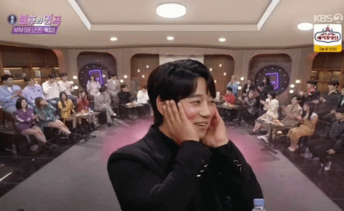 A Man Covering His Ears With His Hands In Front Of A Crowd With A Kbs Logo On The Ceiling GIF