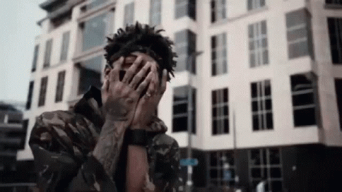 A Man Covering His Face With His Hands In Front Of A Building . GIF