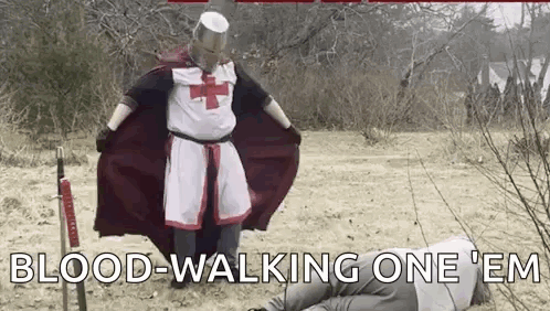A Man Dressed As A Knights Templar Is Standing Next To A Man Laying On The Ground With A Sword . GIF