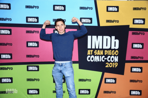 A Man Flexes His Muscles In Front Of A Colorful Backdrop That Says Imdb At San Diego Comic Con 2019 GIF