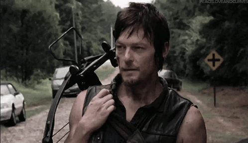 A Man Holding A Bow And Arrow Stands In Front Of A Road Sign That Says Peace Love And Curves GIF