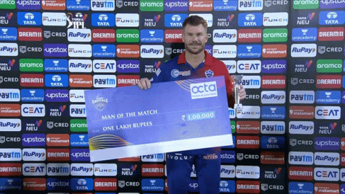 A Man Holding A Cheque In Front Of A Wall With Advertisements For Tata And Ceat GIF