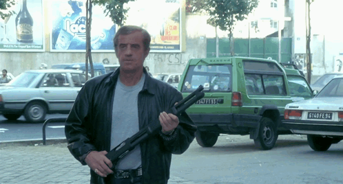 A Man Holding A Gun In Front Of A Green Car With The License Plate R140pe94 GIF