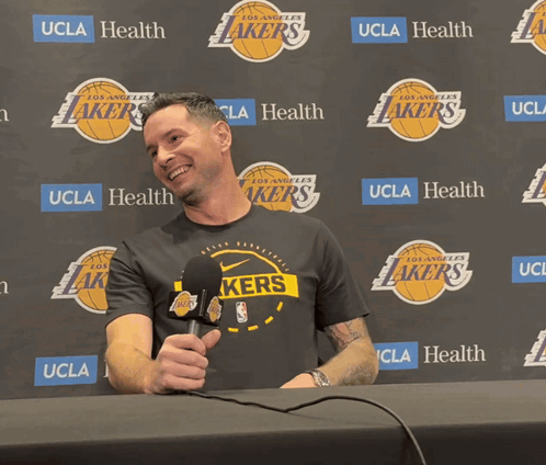A Man Holding A Microphone In Front Of A Wall That Says Lakers GIF
