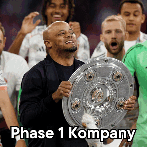A Man Holding A Trophy With The Words " Phase 1 Kompany " On The Bottom GIF