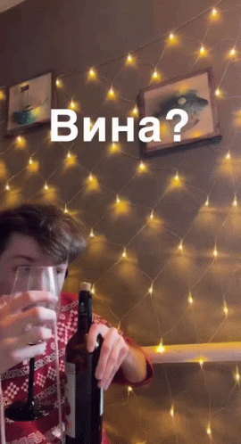 A Man Holds A Bottle Of Wine In Front Of A String Of Lights With The Words Wina Written On It GIF