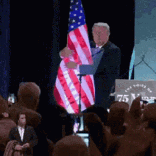 A Man Holds An American Flag In Front Of A Podium That Says 75 GIF