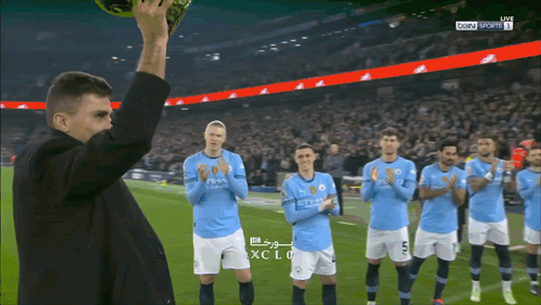 A Man Holds Up A Soccer Ball In Front Of A Group Of Soccer Players Wearing Blue Shirts With The Word City On Them GIF