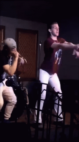 A Man In A 2Pac Shirt Stands Next To Another Man In A Black Shirt GIF