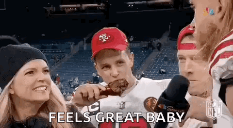 a man in a 49ers hat is eating a piece of meat while a woman holds a child .