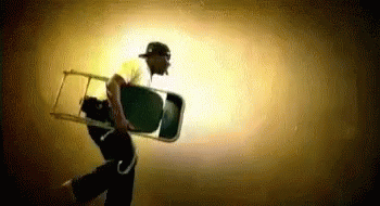 A Man In A Baseball Cap Is Carrying A Chair In A Dark Room . GIF