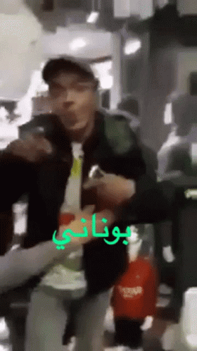 A Man In A Baseball Cap Is Holding A Bottle In Front Of A Sign That Says Botani GIF