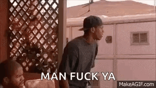 A Man In A Baseball Cap Is Standing In Front Of A Fence With The Words `` Man Fuck Ya '' . GIF