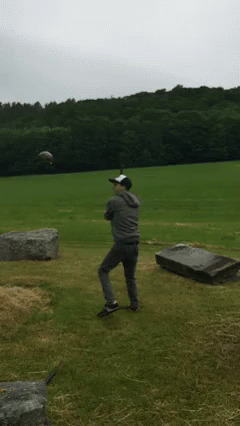 A Man In A Baseball Cap Stands In A Field With A Grave In The Background GIF