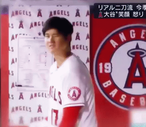 A Man In A Baseball Uniform Is Standing In Front Of A Sign That Says `` Angels '' . GIF