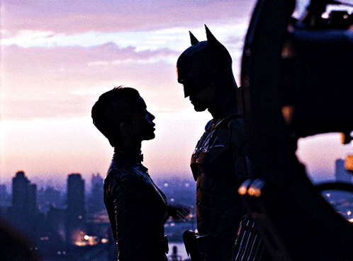 A Man In A Bat Suit Stands Next To A Woman In A Silhouette GIF