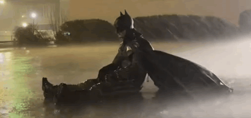 A Man In A Batman Costume Is Sitting On Top Of Another Man In A Puddle Of Water . GIF