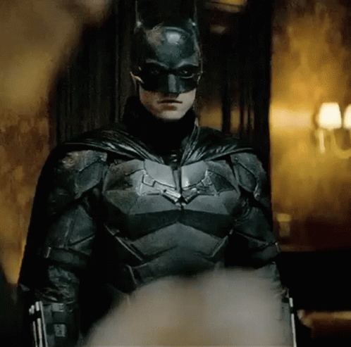A Man In A Batman Costume Is Standing In A Room With A Gun . GIF