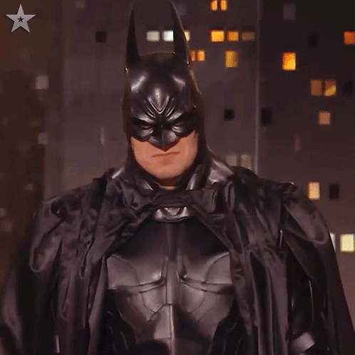 A Man In A Batman Costume Stands In Front Of A Building GIF