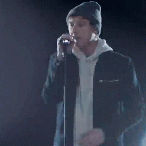 A Man In A Beanie Is Singing Into A Microphone With The Words Our Time Written On It . GIF