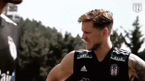 a man in a black adidas tank top is talking to another man in front of a forest .