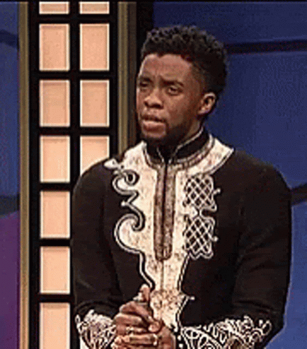 A Man In A Black And White Outfit Is Standing In Front Of A Blue Wall With His Hands Folded . GIF
