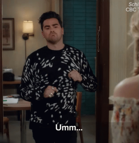 A Man In A Black And White Sweater Stands In Front Of A Door And Says Umm GIF