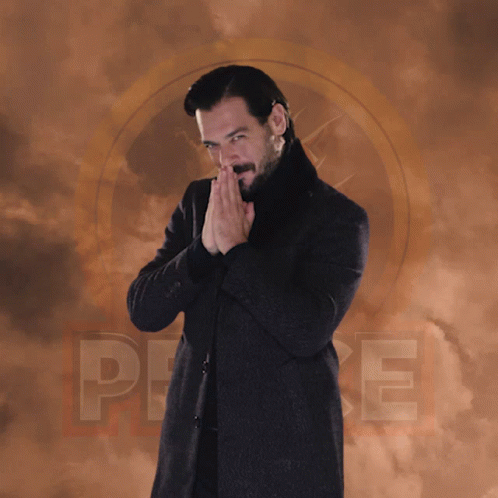 A Man In A Black Coat And Black Turtleneck Stands In Front Of A Cloudy Background With The Letter P On It GIF