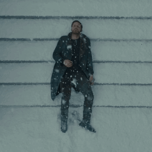 A Man In A Black Coat Is Laying On The Ground In The Snow GIF