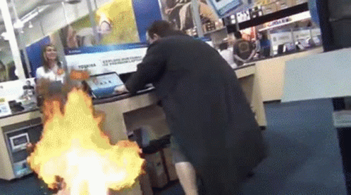 A Man In A Black Coat Is Standing In Front Of A Laptop On Fire GIF