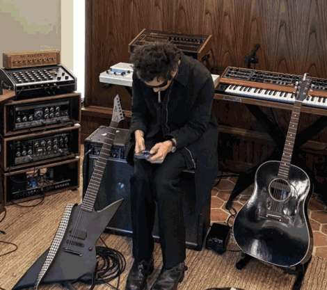 A Man In A Black Coat Sits In Front Of A Keyboard And Guitar GIF