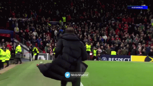 A Man In A Black Coat Stands On A Soccer Field In Front Of A Crowd GIF