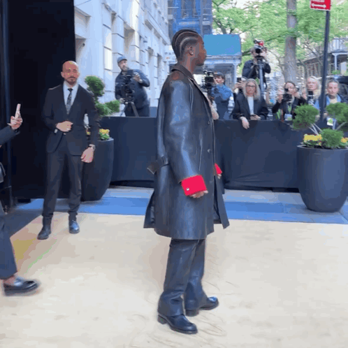 A Man In A Black Coat With Red Cuffs Stands In Front Of A Crowd GIF