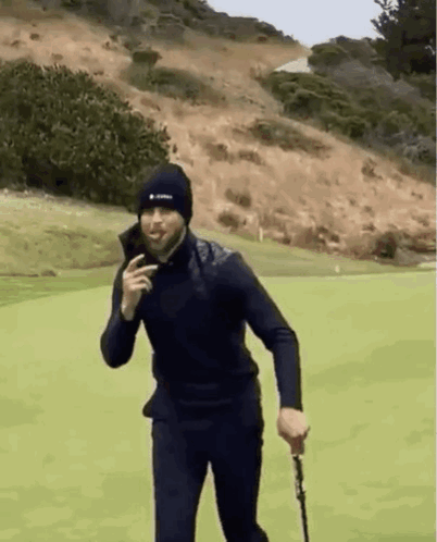 A Man In A Black Hat Is Holding A Golf Club On A Golf Course . GIF