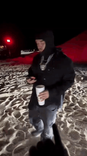 A Man In A Black Hooded Jacket Is Standing On A Beach Holding A Cup . GIF