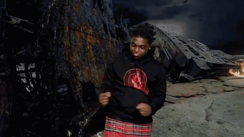 A Man In A Black Hoodie And Plaid Shorts Is Standing In Front Of A Burned Out Building . GIF